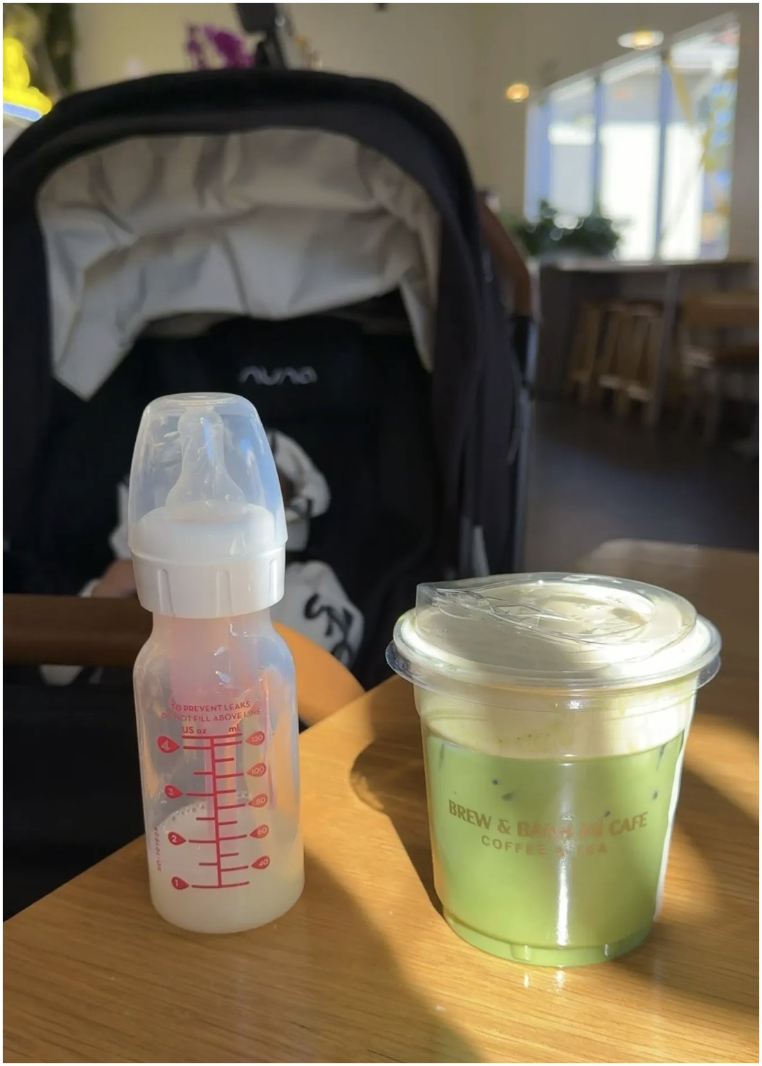 A baby bottle and an iced coffee on a coffee shop table, with an empty stroller behind