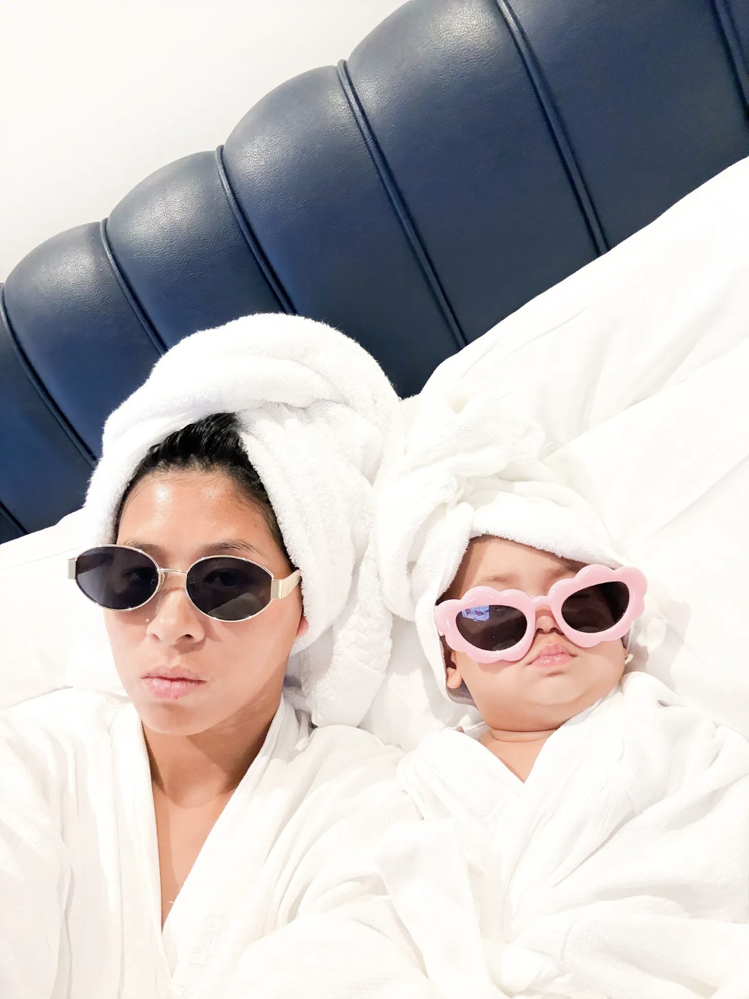 Bethany and her baby lying in bed together in matching white bathrobes, towel hair wraps and sunglasses