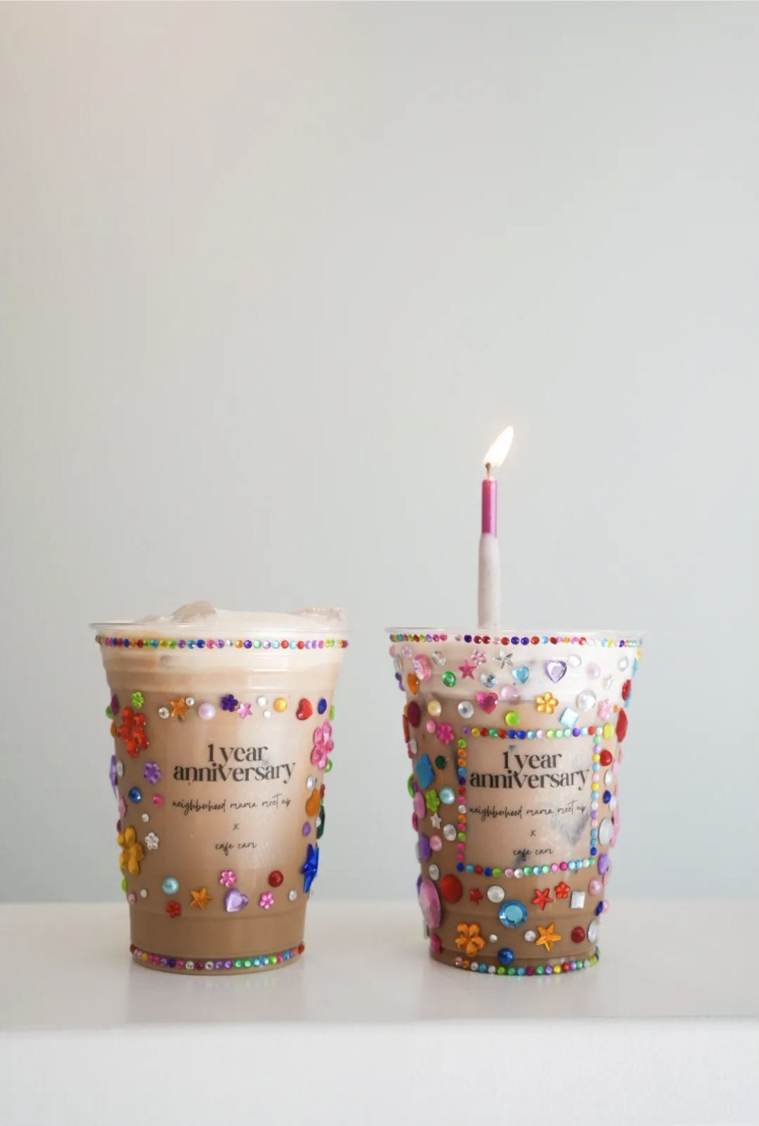 Two iced coffees in rhinestone-covered cups celebrating one year of Neighborhood Mama Meetup, one with a lit candle